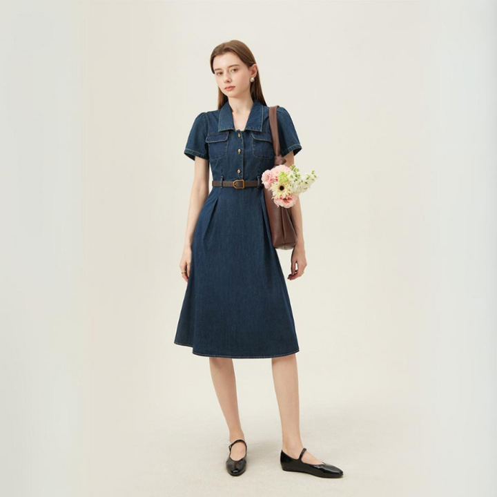 A woman stands against a plain background wearing a belted, short-sleeve denim dress—perfect for summer wear—with black flats. She holds a brown tote bag containing a bouquet of flowers and looks ahead with a neutral expression.