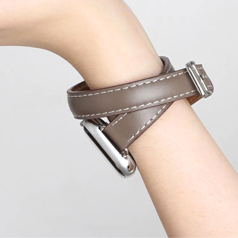 A close-up of a persons wrist wearing a brown genuine leather Apple Watch band with double wrap design, white stitching, and a silver rectangular watch face. The background is plain and light-colored.