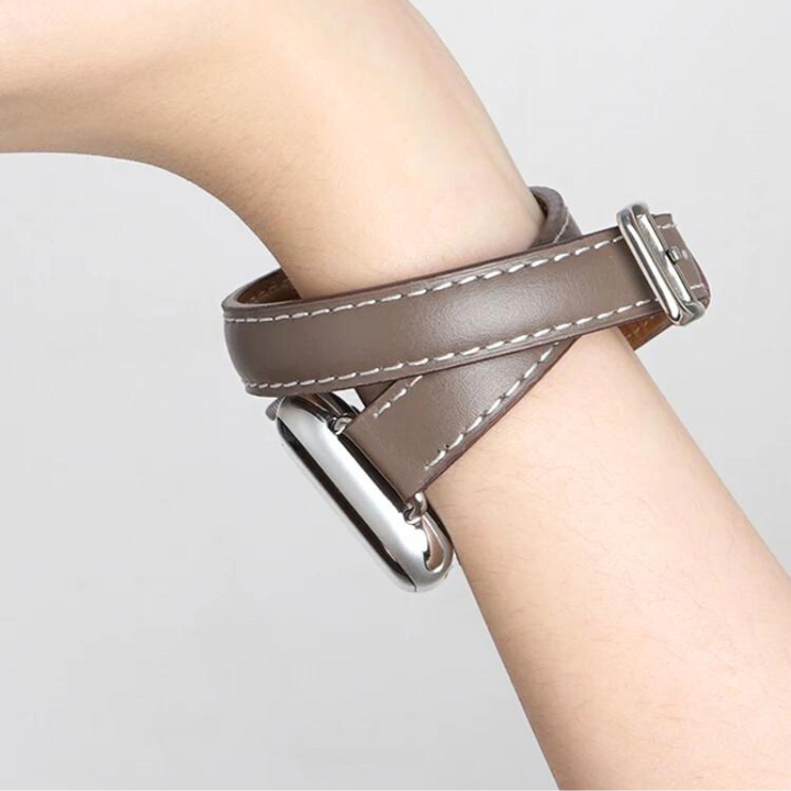 A close-up of a persons wrist wearing a brown genuine leather Apple Watch band with double wrap design, white stitching, and a silver rectangular watch face. The background is plain and light-colored.