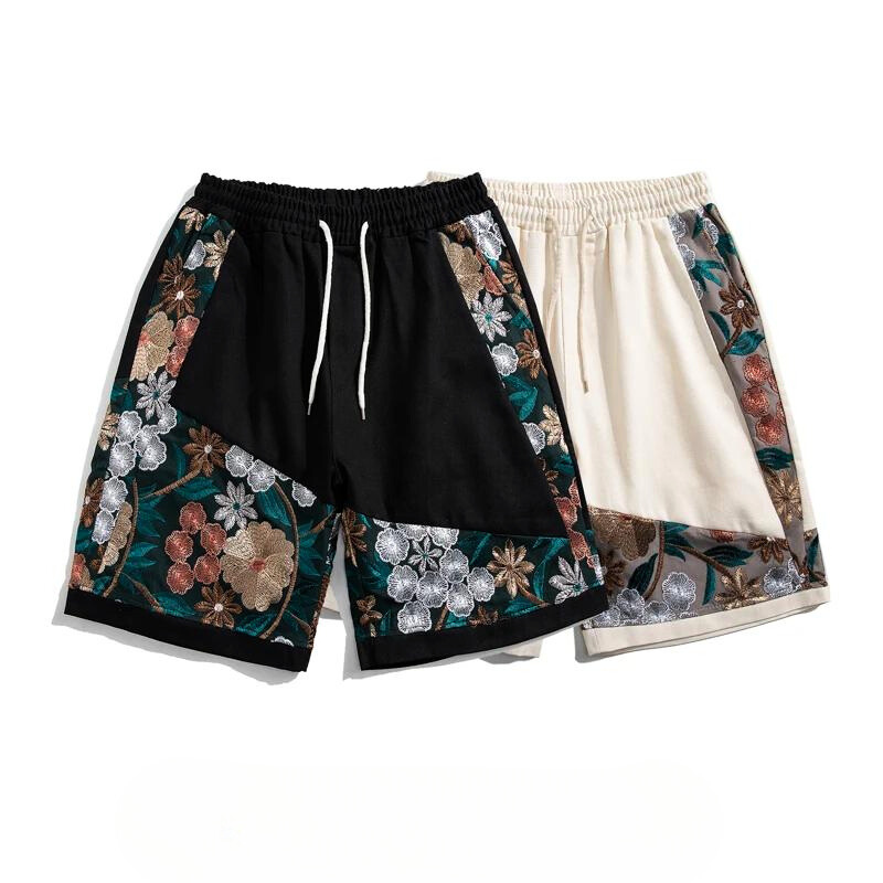 Two pairs of knee-length cotton shorts featuring summer florals embroidered on the sides; one in black with a white drawstring, the other cream with a matching tie. Both are lightweight and adorned with vibrant multicolored flower patterns.