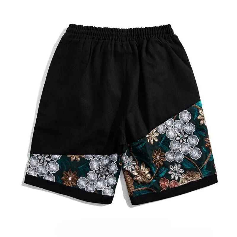 Black, lightweight cotton shorts with elastic waistband featuring decorative panels at the bottom, adorned with intricate summer florals in white, gold, and green embroidery.
