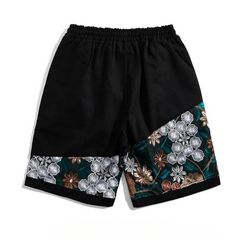 Black, lightweight cotton shorts with elastic waistband featuring decorative panels at the bottom, adorned with intricate summer florals in white, gold, and green embroidery.
