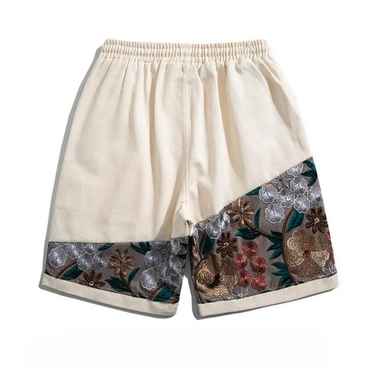 Cream-colored cotton shorts with an elastic waistband, featuring diagonal panels near the hem decorated with intricate summer florals in silver, gold, red, and green tones for a lightweight feel.