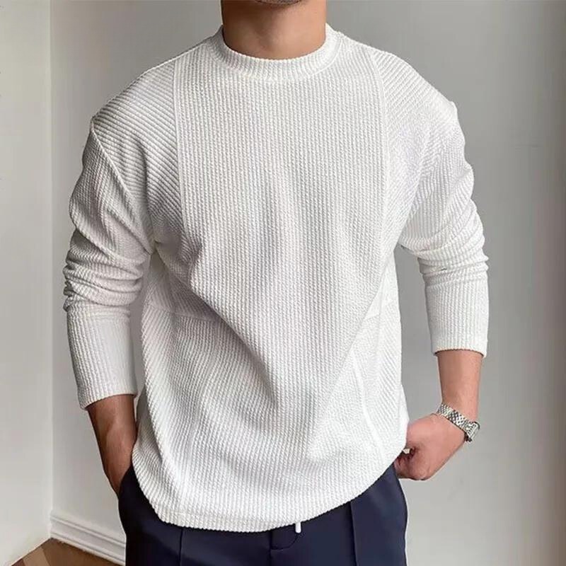 A person wearing a Grayson Crew Classic ribbed knit white long-sleeve sweater and dark pants stands with hands in pockets against a plain light background.