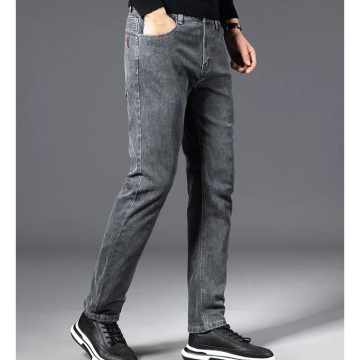 A person stands against a plain gray background, wearing heavyweight denim straight-leg jeans, a black long-sleeve shirt, and black sneakers with white soles. The persons head is not visible in the image.