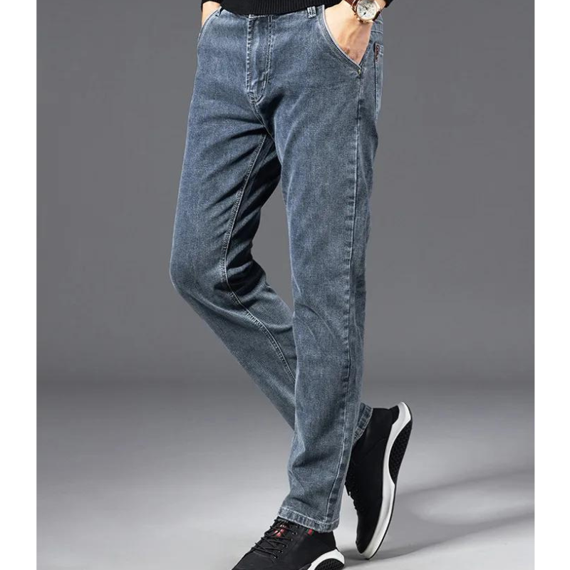 A person wearing straight-leg, blue jeans, a black top, and black athletic shoes stands with hands in pockets against a plain gray background. Only the lower body is visible.