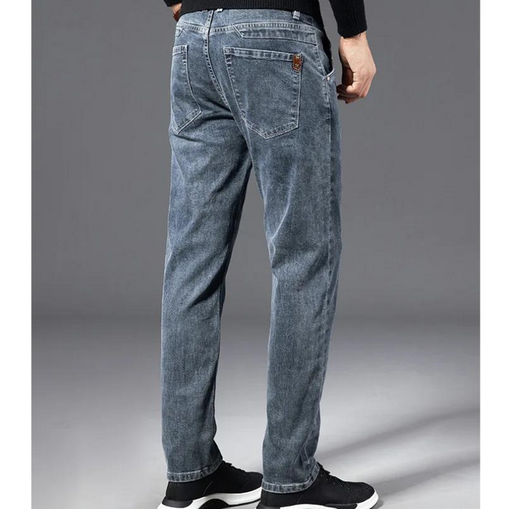 A person wearing faded blue, straight-leg jeans and black sneakers, viewed from the back, standing against a plain gray background.