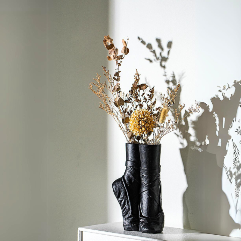 A ballet-inspired vase shaped like black boots holds dried flowers and grasses, casting intricate shadows on a white wall in bright sunlight.