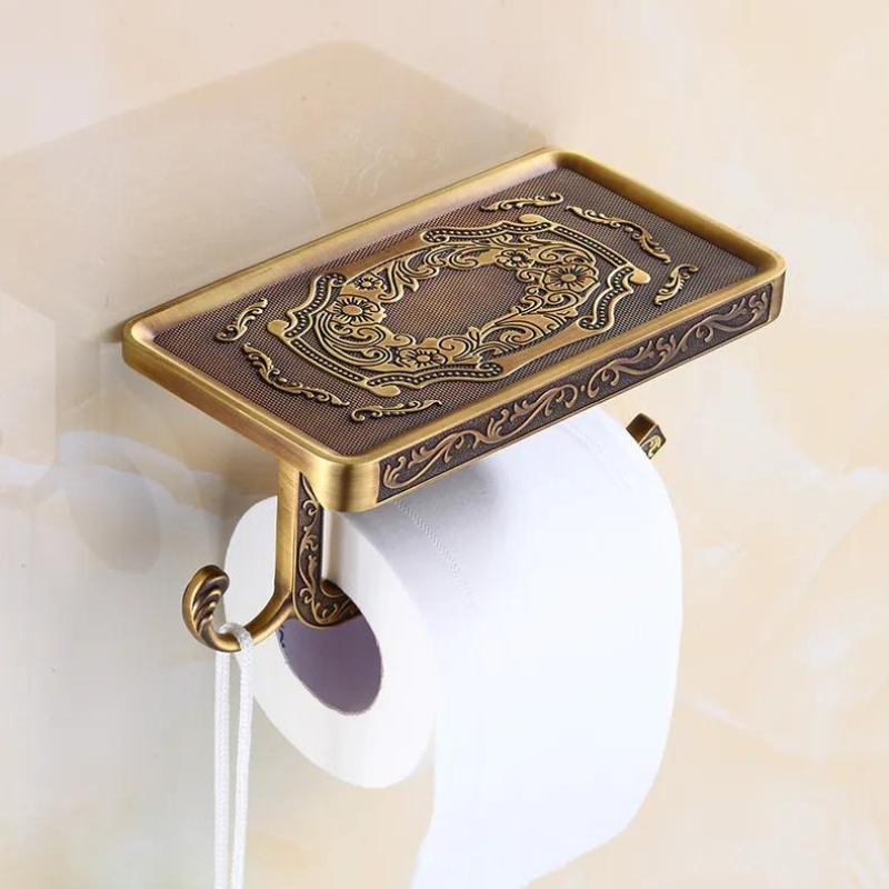 Ornate Victorian toilet paper holder with an antique brass finish, featuring a rectangular decorative tray, roll holder, and hook underneath—an elegant touch to your bathroom decor against a beige wall.