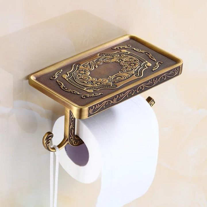 Ornate Victorian toilet paper holder with an antique brass finish, featuring a rectangular decorative tray, roll holder, and hook underneath—an elegant touch to your bathroom decor against a beige wall.