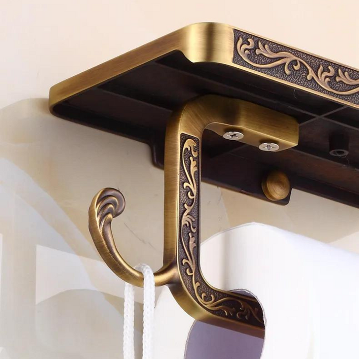 Close-up of an ornate wall hook with an antique brass finish and intricate detailing, mounted on a wall and holding a white cord. The hook, ideal for bathroom decor, features a shelf on top and vintage-style engravings.