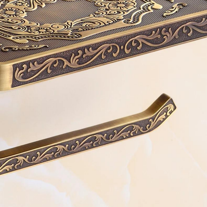 A close-up of an ornate Victorian toilet paper holder with an antique brass finish, featuring intricate, embossed floral patterns and a matching decorative support bar against a light background—perfect for elegant bathroom decor.