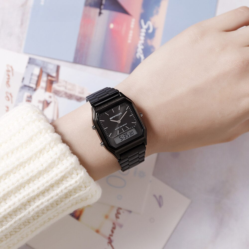 A person wearing a waterproof sports watch with dual display rests their hand on a surface scattered with colorful postcards. The person is dressed in a white knit sweater.