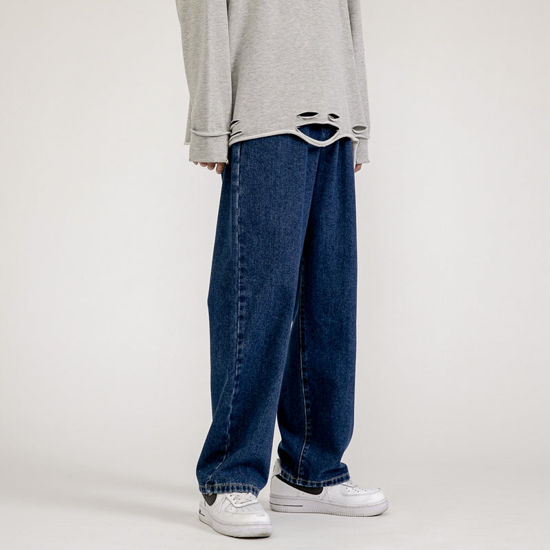 A person wearing a loose, light gray long-sleeve shirt with a distressed hem and men’s baggy jeans in a wide-leg design, paired with white sneakers, channels effortless Korean fashion against a plain background.