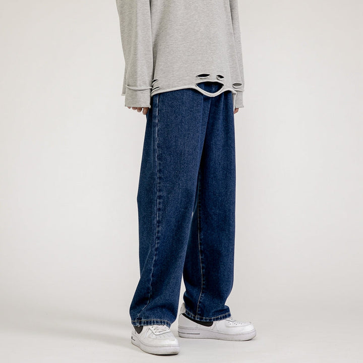 A person wearing a loose, light gray long-sleeve shirt with a distressed hem and men’s baggy jeans in a wide-leg design, paired with white sneakers, channels effortless Korean fashion against a plain background.
