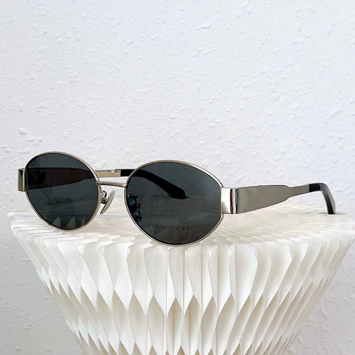 Silver-framed, round black vintage-inspired sunglasses rest on a white, textured, geometric surface against a plain white wall background.