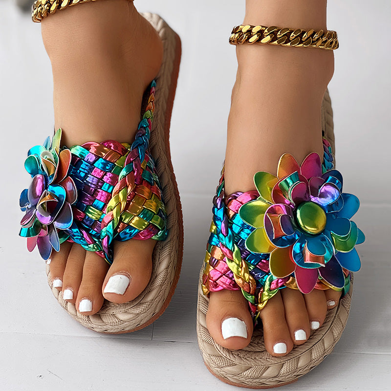 Close-up of feet in colorful, braided women's fashion casual shoes with large, multicolored flower decorations. Toenails are painted white, a gold chain anklet is on one ankle, and the woven sole makes these sandals a perfect vacation essential.