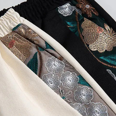 Close-up of three pieces of fabric—cream, taupe, and black—each with intricate summer florals embroidered in metallic and colored threads. These lightweight fabrics have elastic, gathered waistbands.