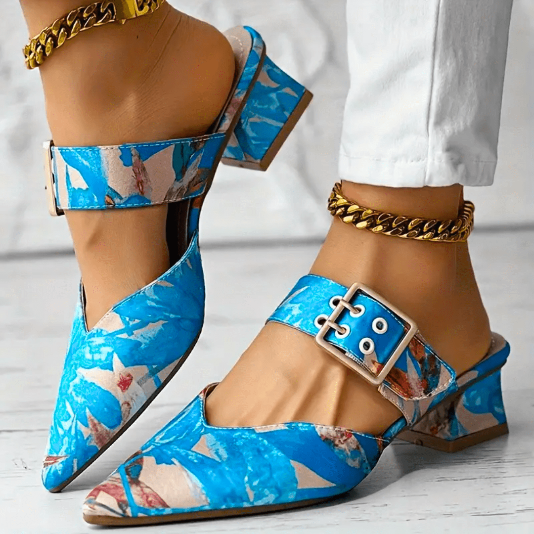 A person wearing blue patterned pointed toe mules with buckle straps and block heels, paired with gold chain anklets and white pants on a light-colored floor.