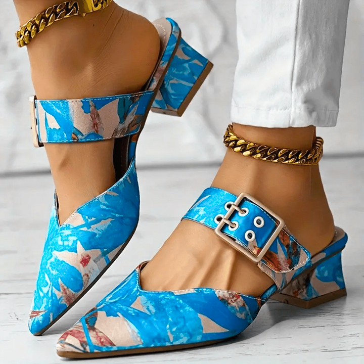 A person wearing blue patterned pointed toe mules with buckle straps and block heels, paired with gold chain anklets and white pants on a light-colored floor.