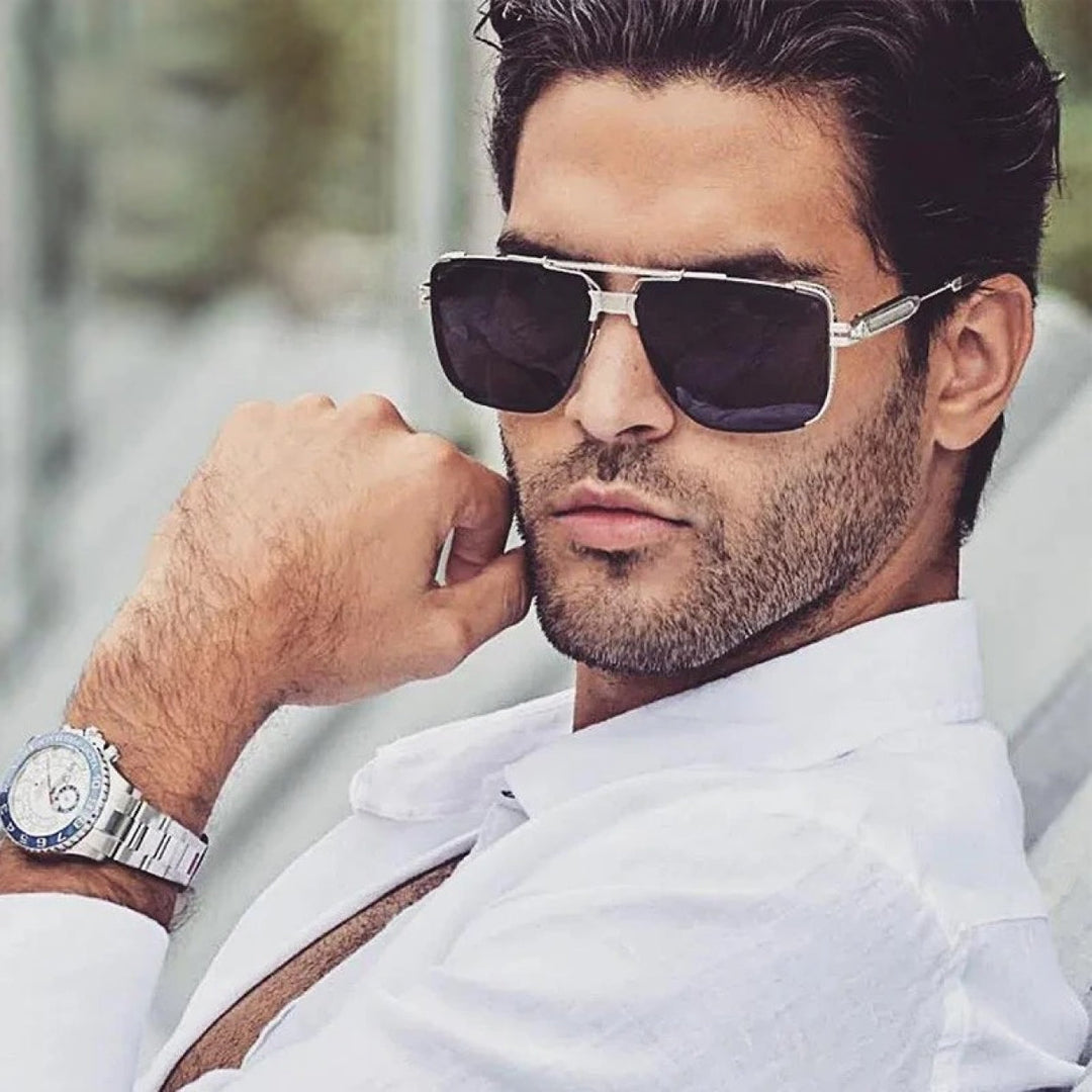 A man with short dark hair wears vintage sunglasses, a white shirt, and a silver watch, looking confidently at the camera with his hand resting near his face.