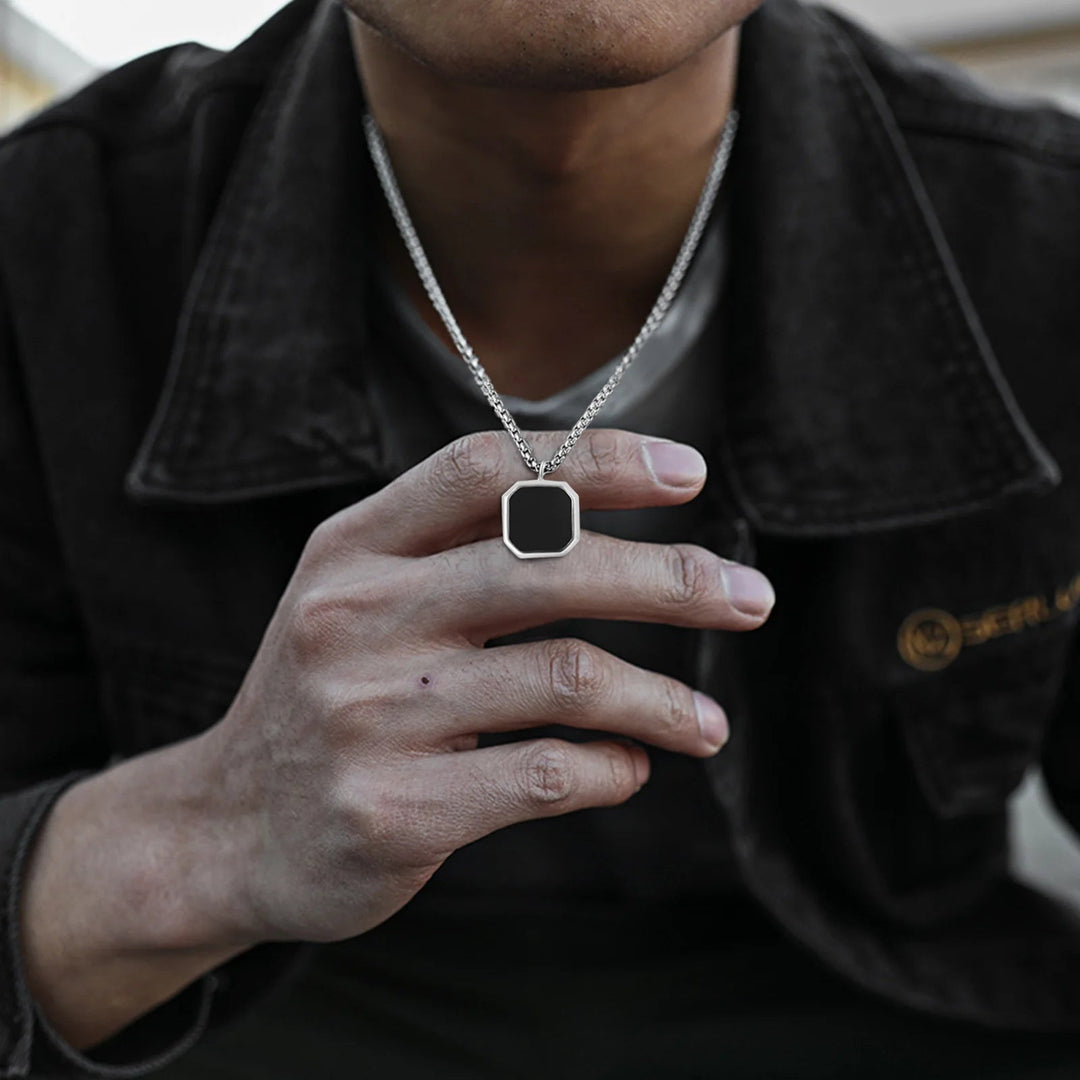 A person wearing a dark jacket holds a silver pendant necklace with a blank, black, octagonal charm resembling an Onyx Stone Amulet close to their chest. Their face is partially out of frame, highlighting the collectable jewelry.