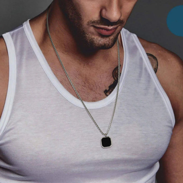 A man wearing a white sleeveless shirt and a silver chain necklace with a square Onyx Stone Amulet; part of his tattoo is visible on his upper chest, adding to the allure of this collectable jewelry piece.