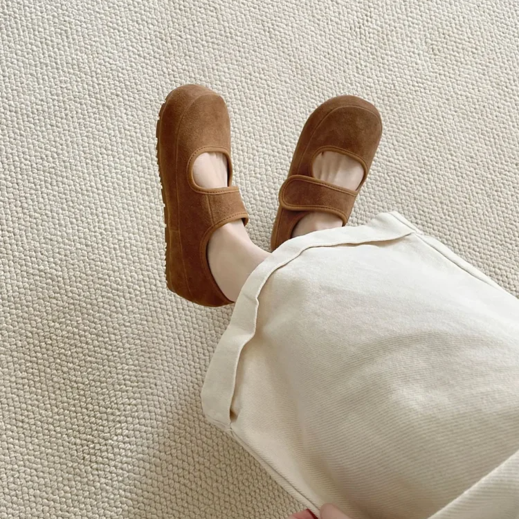 A person wearing a cream-colored skirt and brown suede Mary Janes with a round toe stands on a textured, light-colored carpet, with one hand holding the skirt.