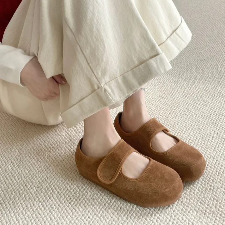 A person wearing cream-colored wide-leg pants and a red and white top sits on a carpeted floor, showing their feet in brown suede Mary Janes with velcro straps and a cushioned insole for added comfort.