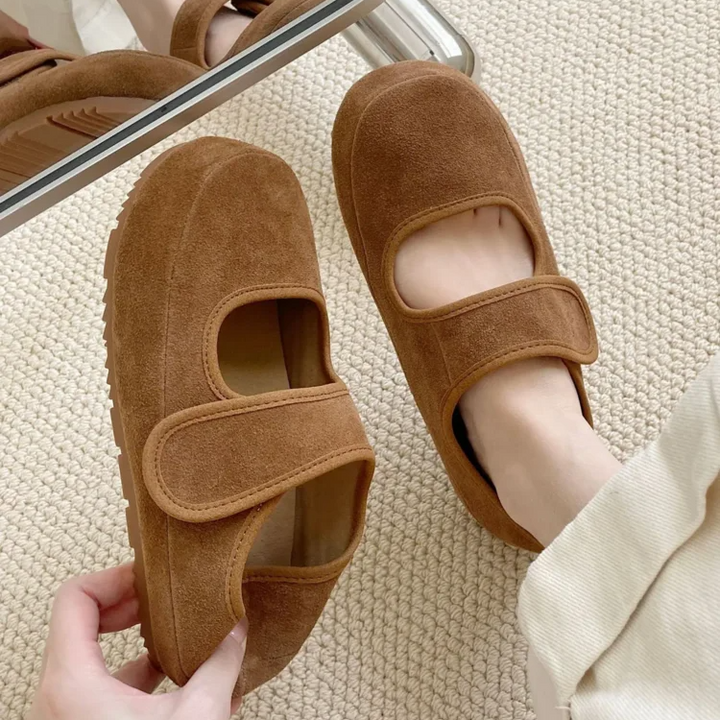 A person wearing cream-colored pants tries on a pair of brown suede Mary Janes with Velcro straps and a cushioned insole, sitting on a light textured carpet near a mirror.