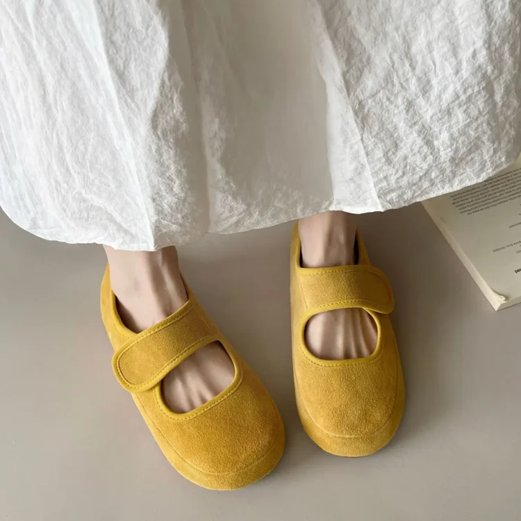A person wearing a long, white, crinkled skirt stands on a light surface, showcasing yellow suede Mary Janes with velcro straps and a cushioned insole. A partially visible book lies nearby.