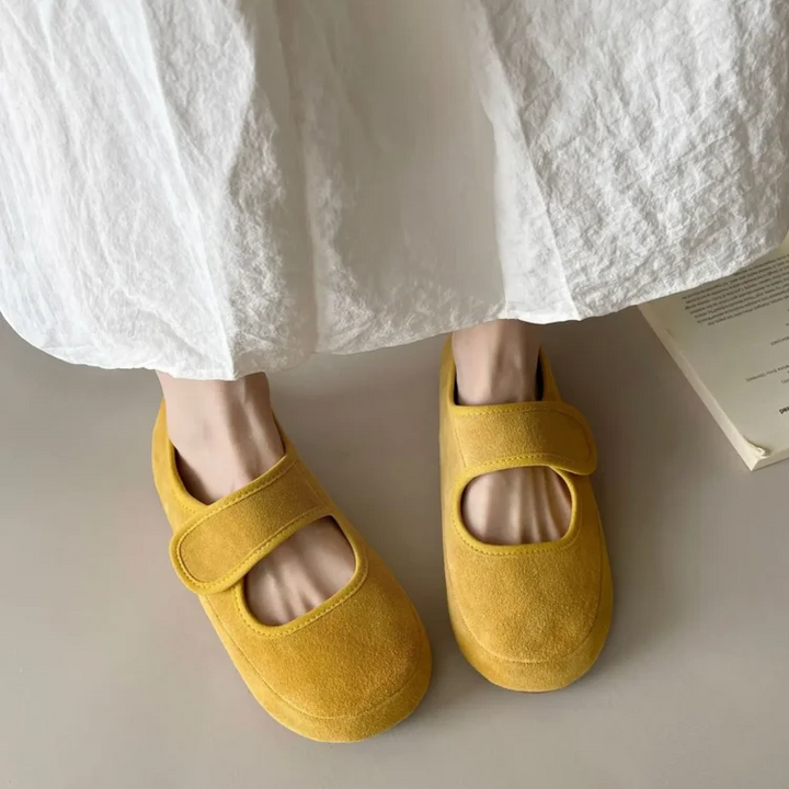 A person wearing a long, white, crinkled skirt stands on a light surface, showcasing yellow suede Mary Janes with velcro straps and a cushioned insole. A partially visible book lies nearby.