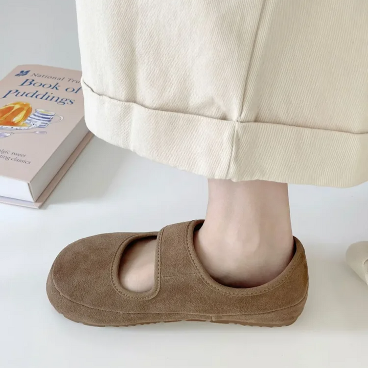 A person wearing beige pants and brown suede Mary Janes with a cushioned insole stands near a book on the floor.