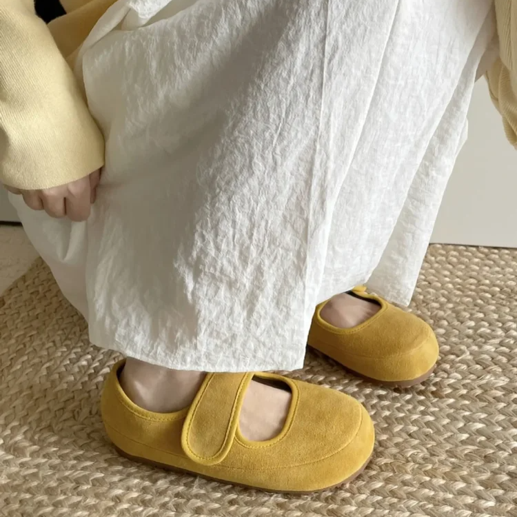 A person in a long white skirt and light yellow sweater crouches on a woven mat, showing off yellow Mary Janes with a round toe and cushioned insole.