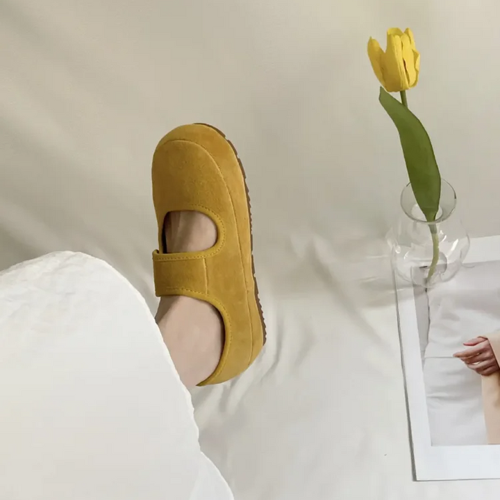A person wearing a yellow round toe Mary Jane shoe rests their foot on a white surface near a glass vase with a yellow tulip and a photo of a hand. The scene has a soft, minimalistic aesthetic.