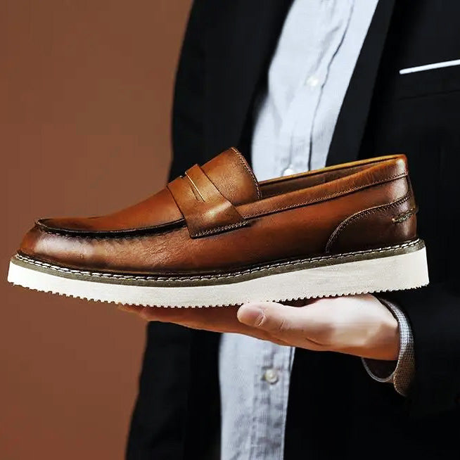 A person in a suit jacket and dress shirt holds a brown Italian leather penny loafer with a white sole, displaying the elegant business casual shoe against a plain brown background.