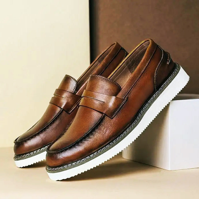 A pair of brown Italian leather penny loafers with white, textured rubber soles are displayed, one resting on a white box and the other on a beige surface, against a two-tone beige and brown background—perfect for business casual style.