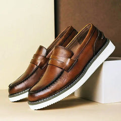 A pair of brown Italian leather penny loafers with white, textured rubber soles are displayed, one resting on a white box and the other on a beige surface, against a two-tone beige and brown background—perfect for business casual style.