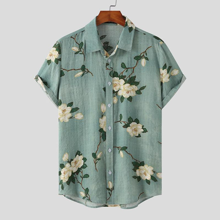 Short-sleeved, light teal Mens Hawaiian Shirt featuring white flower prints and green leaves, displayed on a wooden hanger against a plain light gray background.