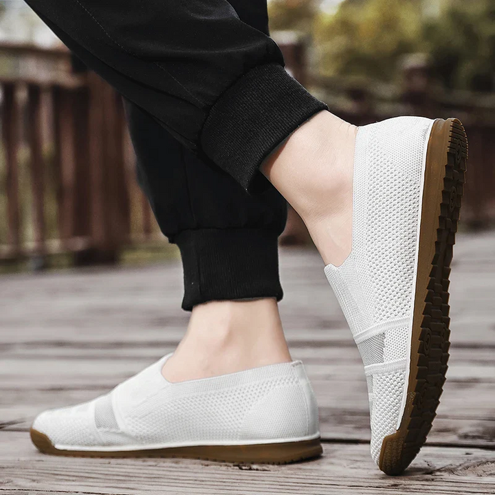 A person wearing lightweight slip-on mesh shoes with brown soles stands on a wooden surface outdoors. The black pants complement the shoes, while blurred greenery and a fence appear in the background.