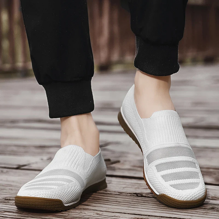 A person wearing lightweight slip-on sneakers with a light brown sole stands on a wooden surface. The white shoes pair with black cuffed pants, showcasing the effortless style and comfort of breathable knit shoes.