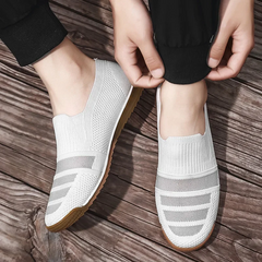 A person wearing lightweight slip-on knit sneakers with light brown soles is adjusting the cuff of their black pants while standing on a wooden surface.