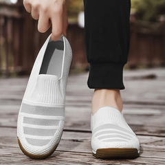 A person wearing black pants is putting on a white lightweight slip-on shoe with a brown sole while standing outdoors on a wooden surface. The other foot is already wearing a matching shoe.