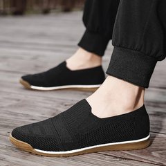 A person wearing black lightweight slip-on sneakers with white midsoles, brown outsoles, and a cushioned insole, paired with black jogger pants, standing on a wooden surface.