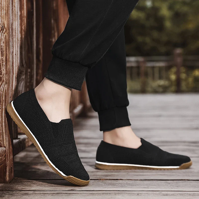 A person wearing black slip-on shoes with a breathable knit upper and white soles is standing on a wooden deck. The shoes feature tan bottoms, and the person is dressed in black pants with trees visible in the background.