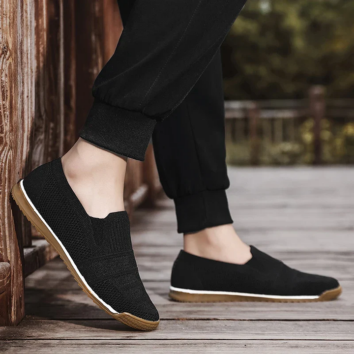 A person wearing black slip-on shoes with a breathable knit upper and white soles is standing on a wooden deck. The shoes feature tan bottoms, and the person is dressed in black pants with trees visible in the background.