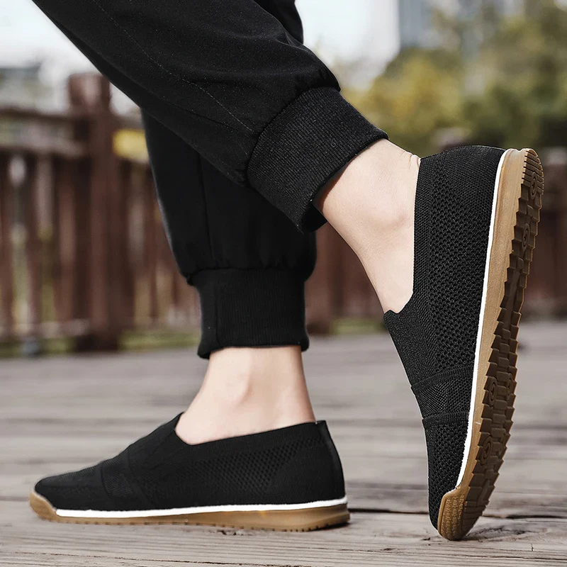A person wearing black lightweight slip-on casual shoes with light brown soles stands on a wooden surface outdoors. The cushioned insole offers comfort, while black pants and a slightly blurred background complete the scene.
