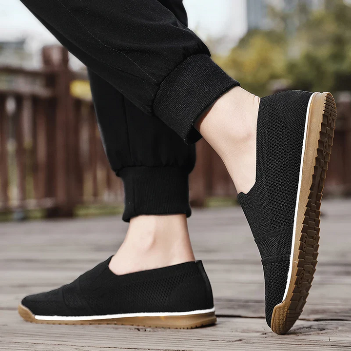 A person wearing black lightweight slip-on casual shoes with light brown soles stands on a wooden surface outdoors. The cushioned insole offers comfort, while black pants and a slightly blurred background complete the scene.