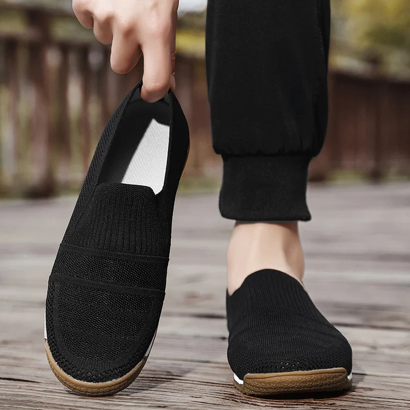 A person wearing lightweight slip-on shoes stands on a wooden surface, lifting one shoe slightly while dressed in black pants. The image focuses on the shoes’ cushioned insole and the lower legs.