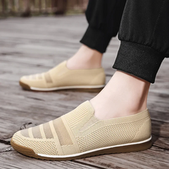 A person wearing lightweight slip-on mesh sneakers with cushioned insoles and brown soles stands on a wooden surface, their legs in black pants visible.